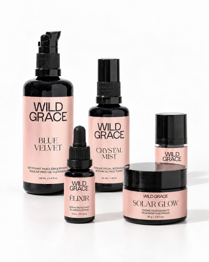 A set of five Wild Grace skincare bottles and jars arranged on a white background, featuring black packaging with soft pink labels. The products include Blue Velvet, Crystal Mist, Nova, Elixir, and Solar Glow.