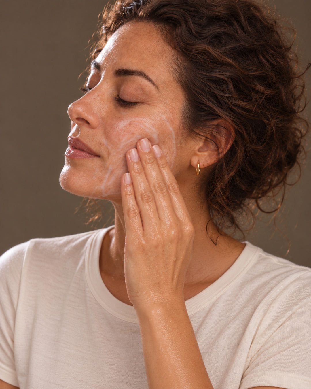 A woman with curly brown hair applies a white skincare cream to her cheek. She is wearing a simple white t-shirt and a small gold hoop earring, set against a solid neutral brown background.