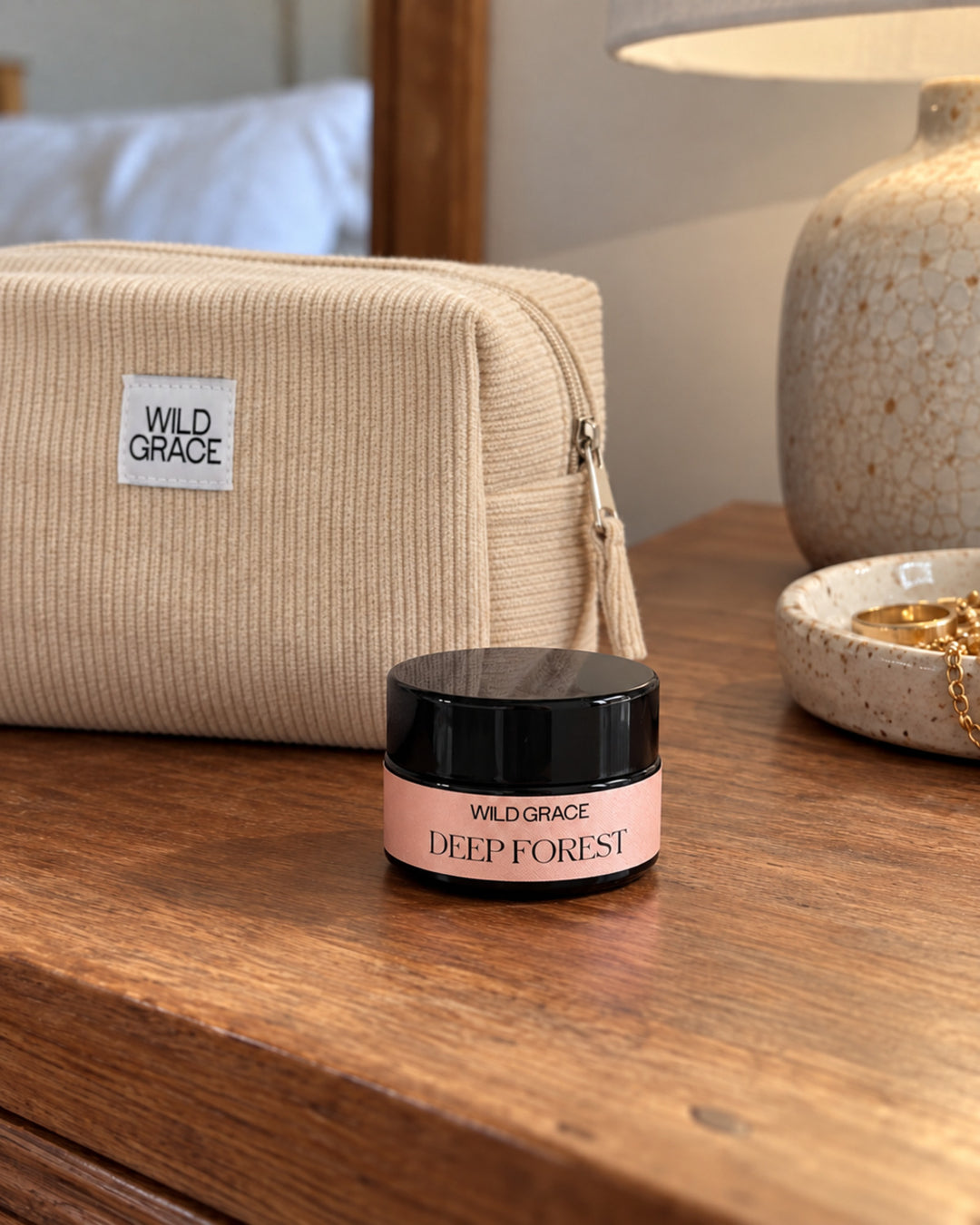 A small Wild Grace Deep Forest cosmetic jar with a black lid and pink label sits on a wooden table, with a matching beige ribbed zip pouch and a lamp in the background.