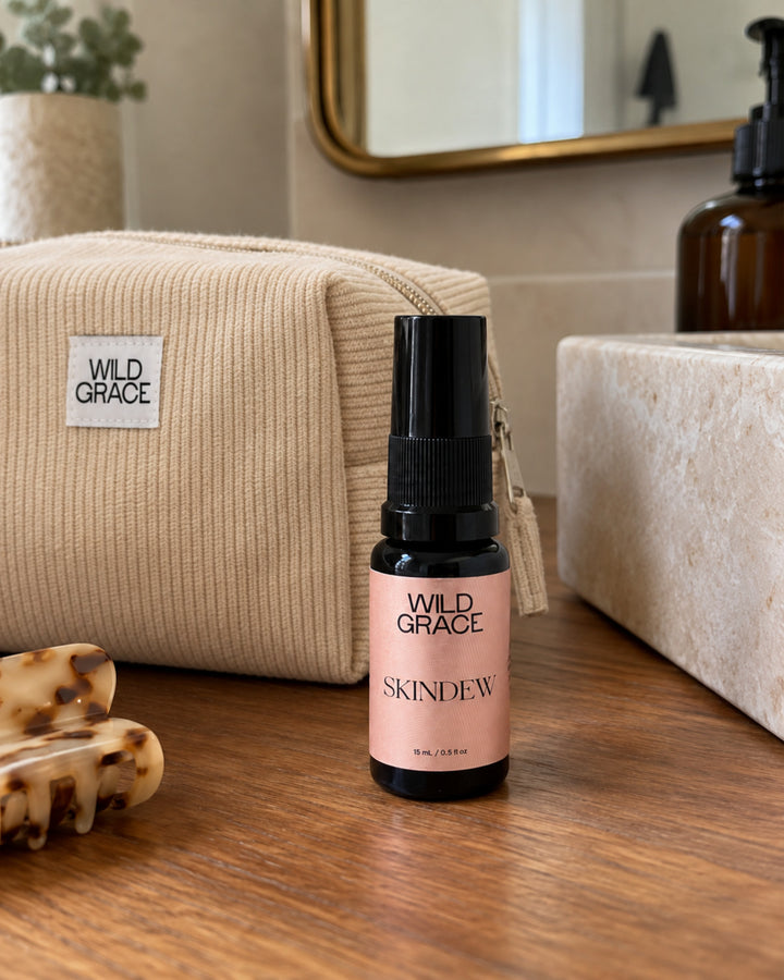 A small amber spray bottle labeled WILD GRACE SKINDEW stands on a wooden surface in front of a beige corduroy zip pouch with a WILD GRACE label. The scene includes a stone sink, mirror, and soap dispenser in a bathroom setting.