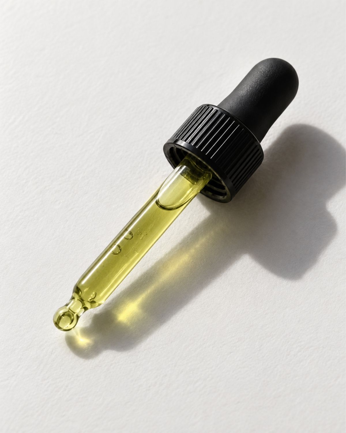 A glass dropper bottle filled with pale yellow oil, featuring a black ribbed cap and rubber bulb, photographed on a light gray textured background with a soft shadow.