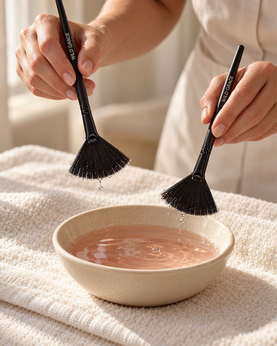 Esthetician holding two black WILD GRACE fan brushes above a ceramic bowl of facial tonic.