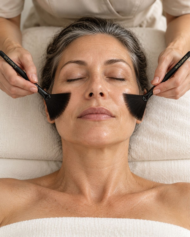 Esthetician applying facial tonic with two WILD GRACE fan brushes, one on each cheek, during a professional facial.