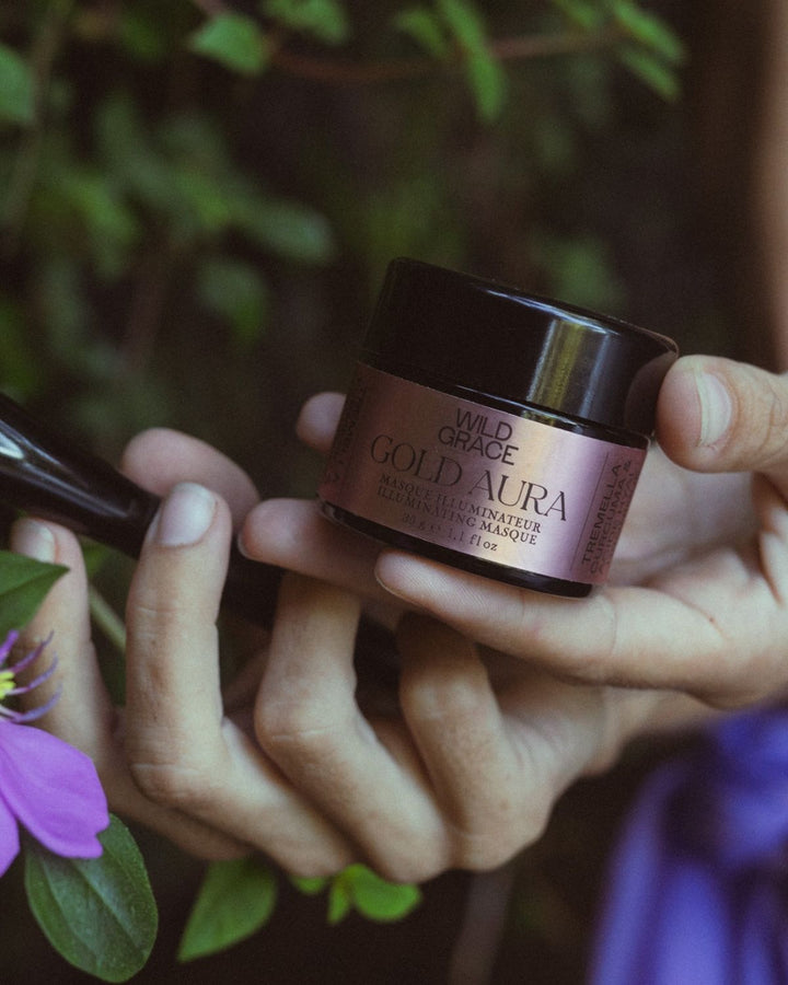 jar of gold aura illuminating mask by wild grace theld in hands of woman with green nature background
