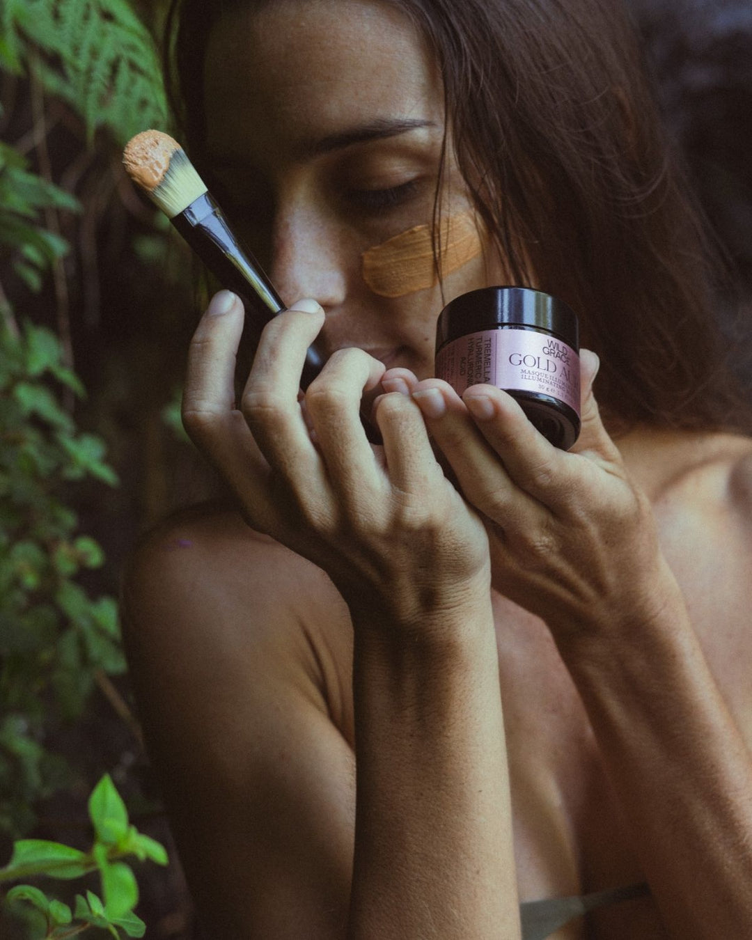 Woman smrlling the gold aura illuminating mask by wild grace holding the jar of the same product in front of her.