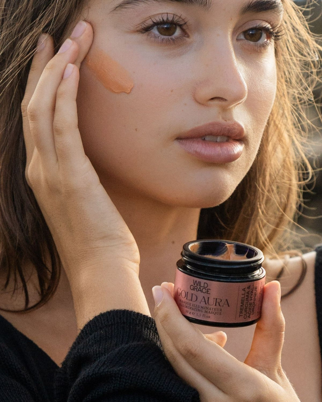 Woman applying the gold aura illuminating mask by wild grace to her face with a jar of the same product in front of her.