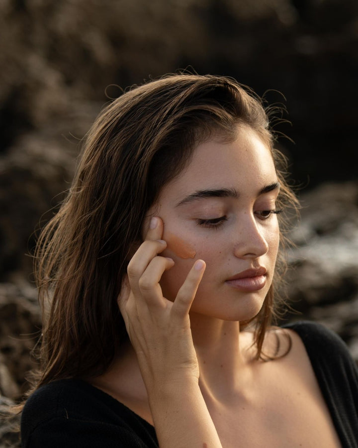 Woman applying the gold aura illuminating mask by wild grace to her face with a jar of the same product in front of her.