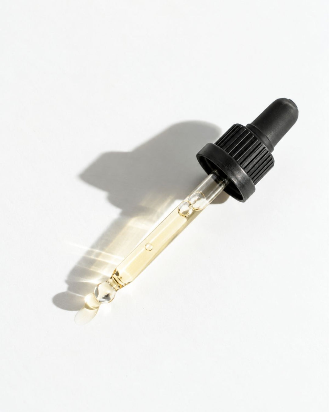 A clear glass dropper bottle filled with pale golden liquid, featuring a black ribbed dropper cap, photographed on a clean white background with a soft shadow.