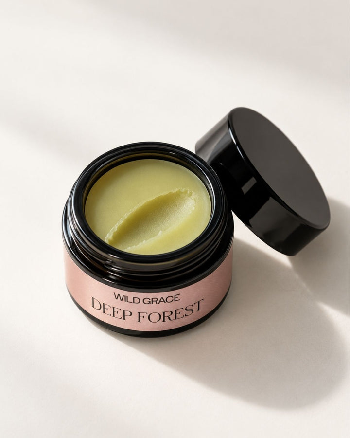 An open jar of Wild Grace Deep Forest skincare cream showing a pale yellow-green balm inside, with its black lid resting beside it on a clean white background.