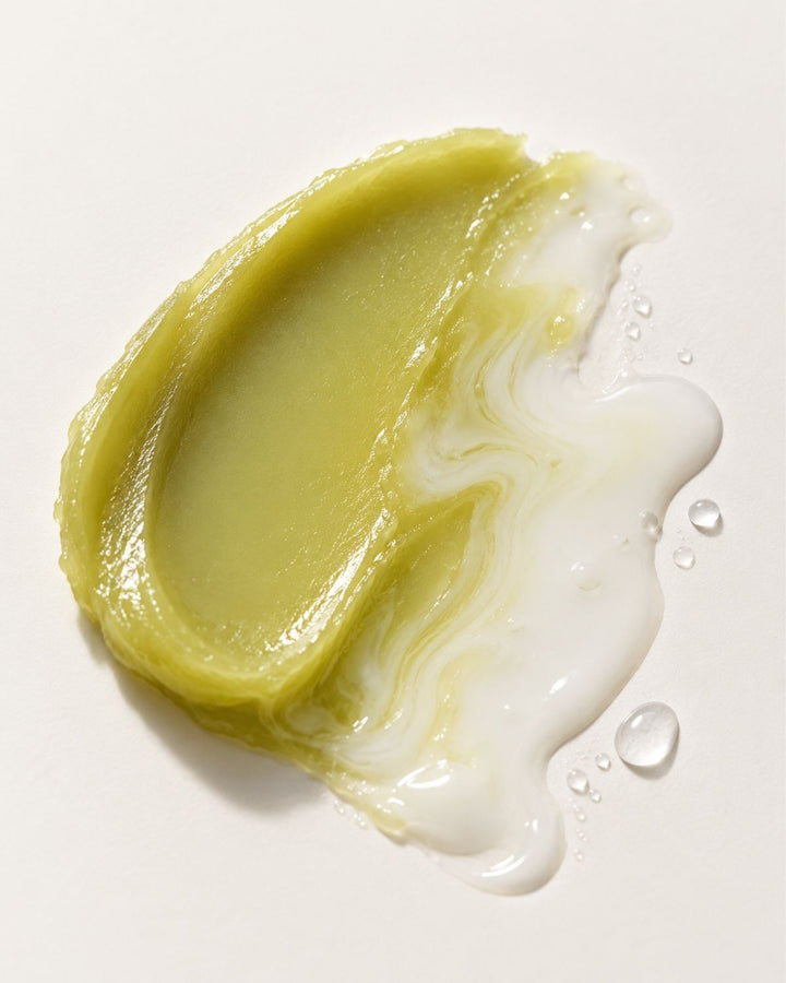 A dollop of creamy, pale yellow-green cosmetic cream or balm with a glossy finish, spread on a white surface with a translucent white liquid swirled around it and small droplets nearby.