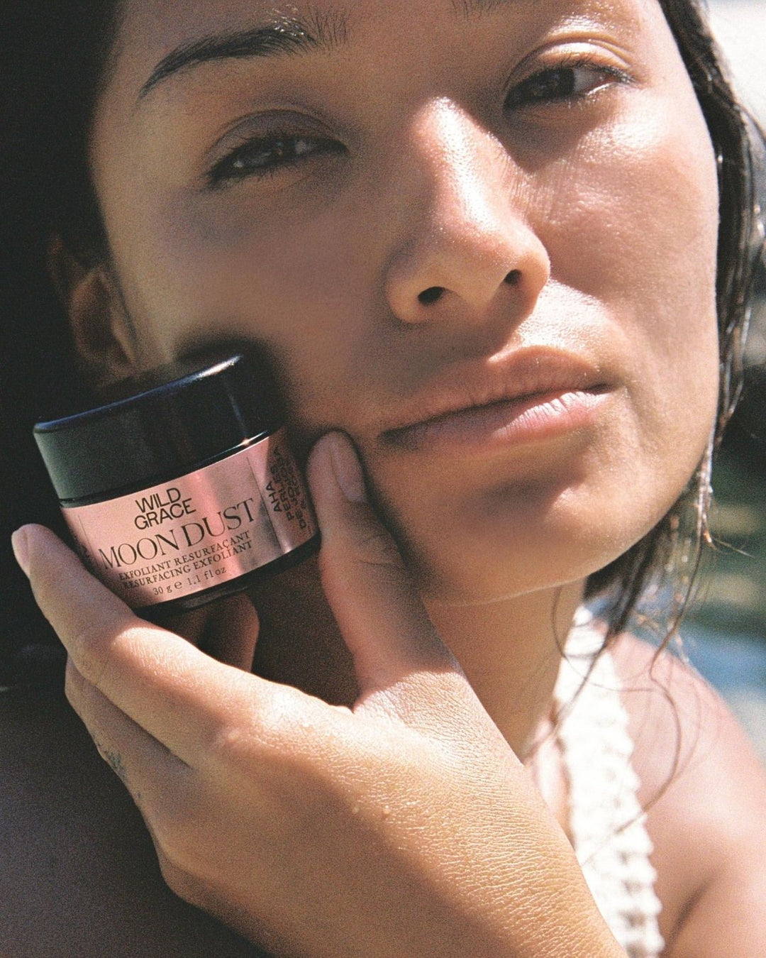 woman holding a jar of moon dust exfoliant by wild grace. sunlight is shining on her face