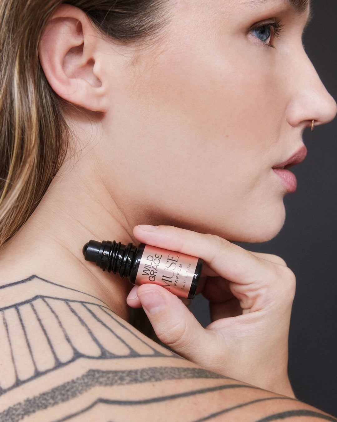 Woman with a large tattoo on her back holding a bottle of MUSE perfume oil by wild grace close to her neck