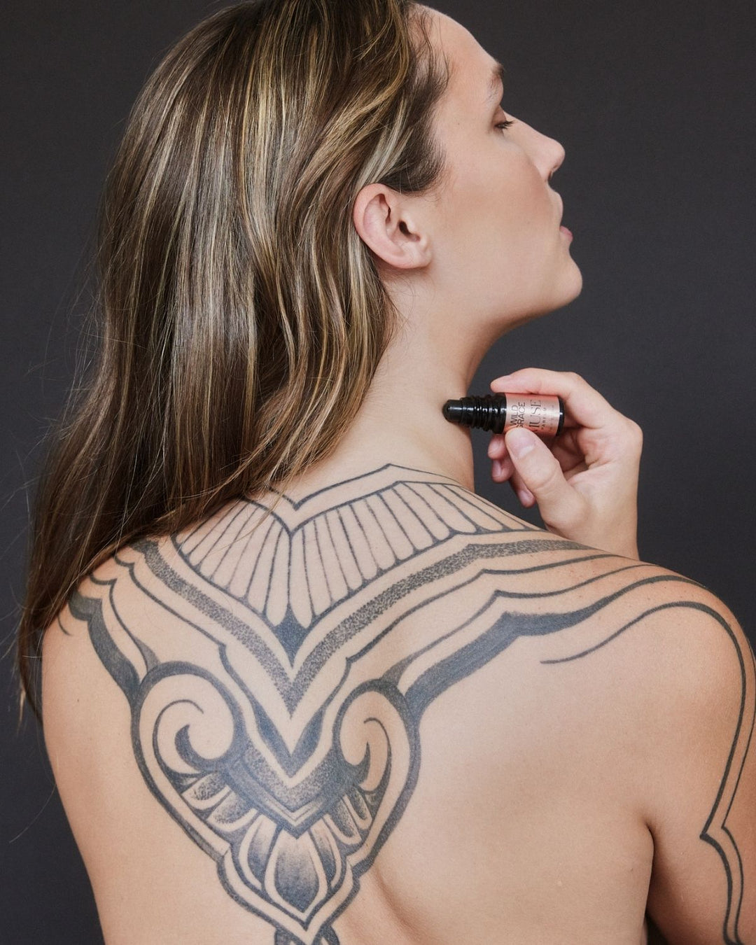 Woman with a large tattoo on her back holding a bottle of MUSE perfume oil by wild grace against a dark background