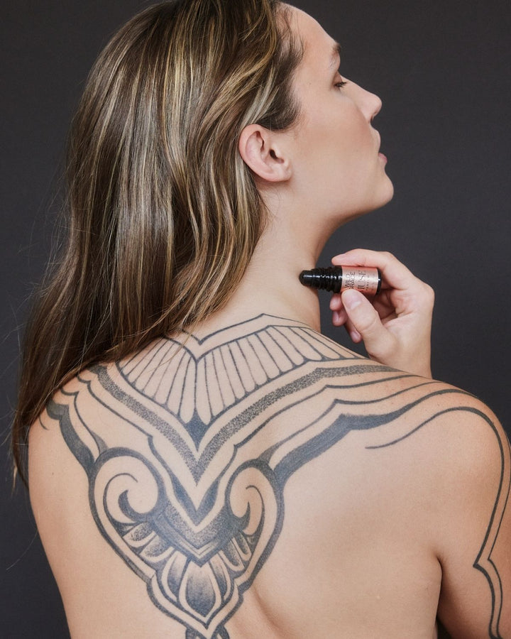Woman with a large tattoo on her back holding a bottle of MUSE perfume oil by wild grace against a dark background