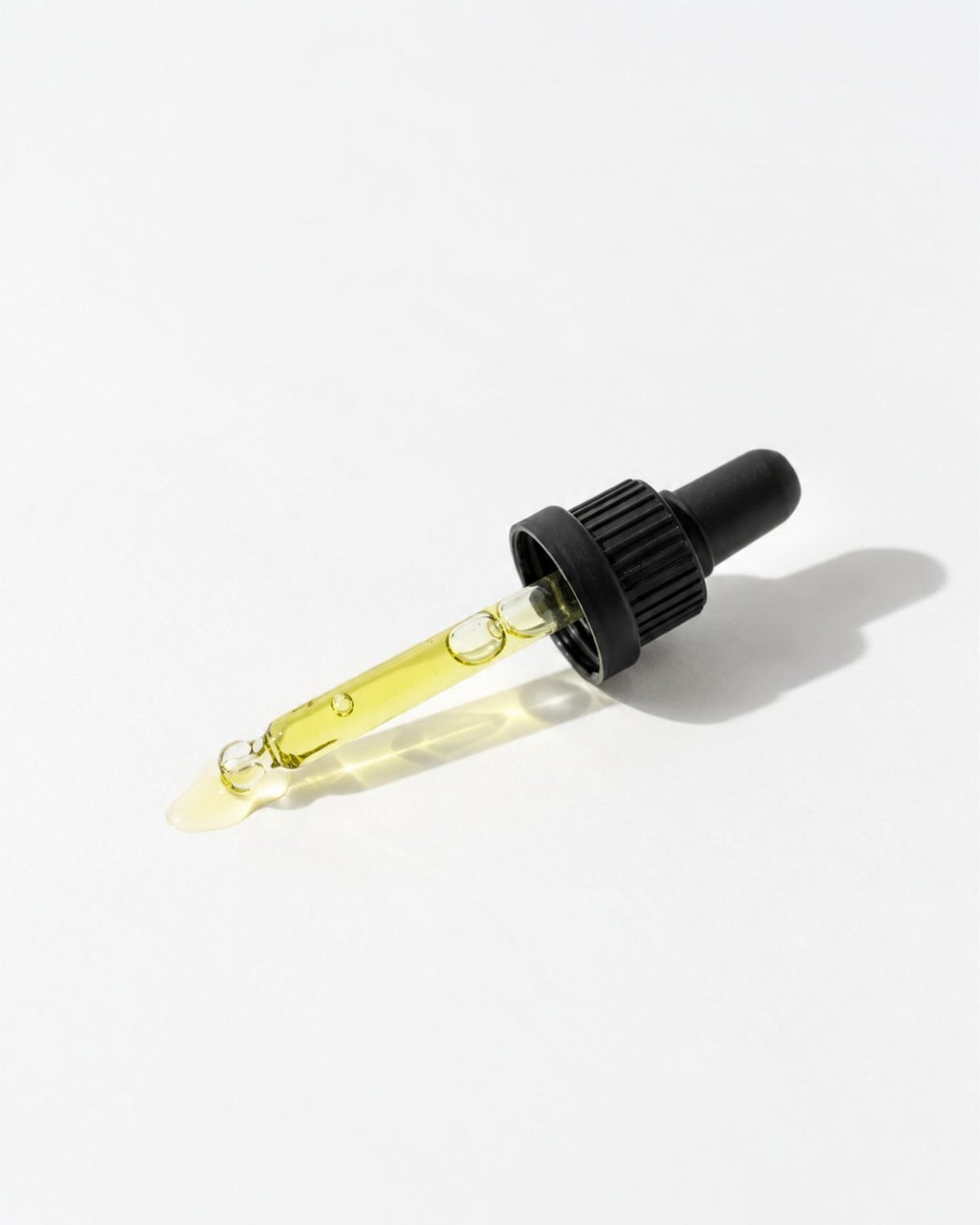A glass dropper bottle filled with pale yellow oil, featuring a black ribbed dropper cap, photographed on a clean white background with a soft shadow.