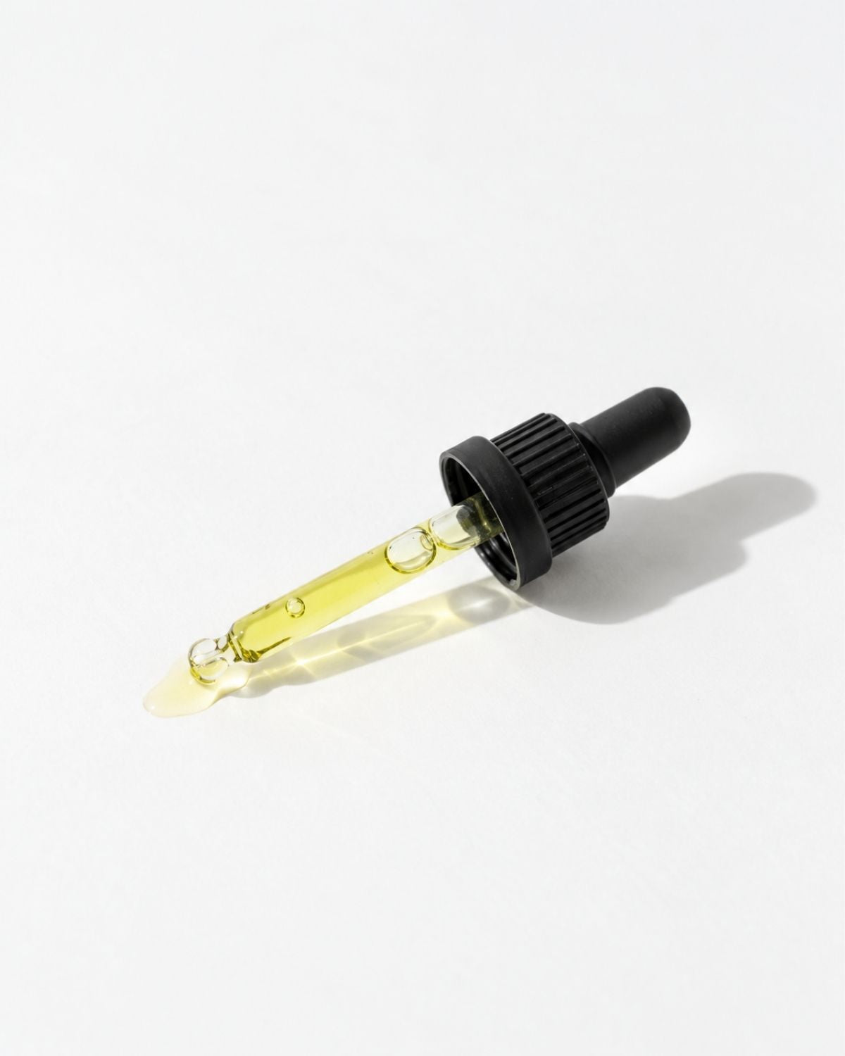 A glass dropper bottle filled with pale yellow oil, featuring a black ribbed dropper cap, photographed on a clean white background with a soft shadow.