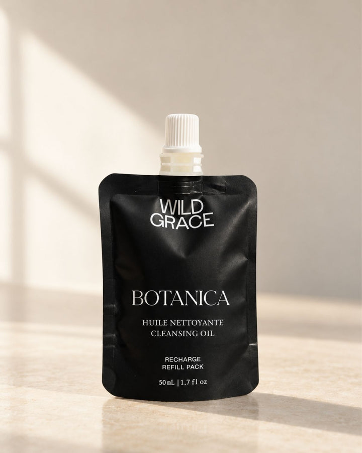 A black refill pouch of Wild Grace Botanica Cleansing Oil with a white cap, set against a warm beige background with soft window shadows.