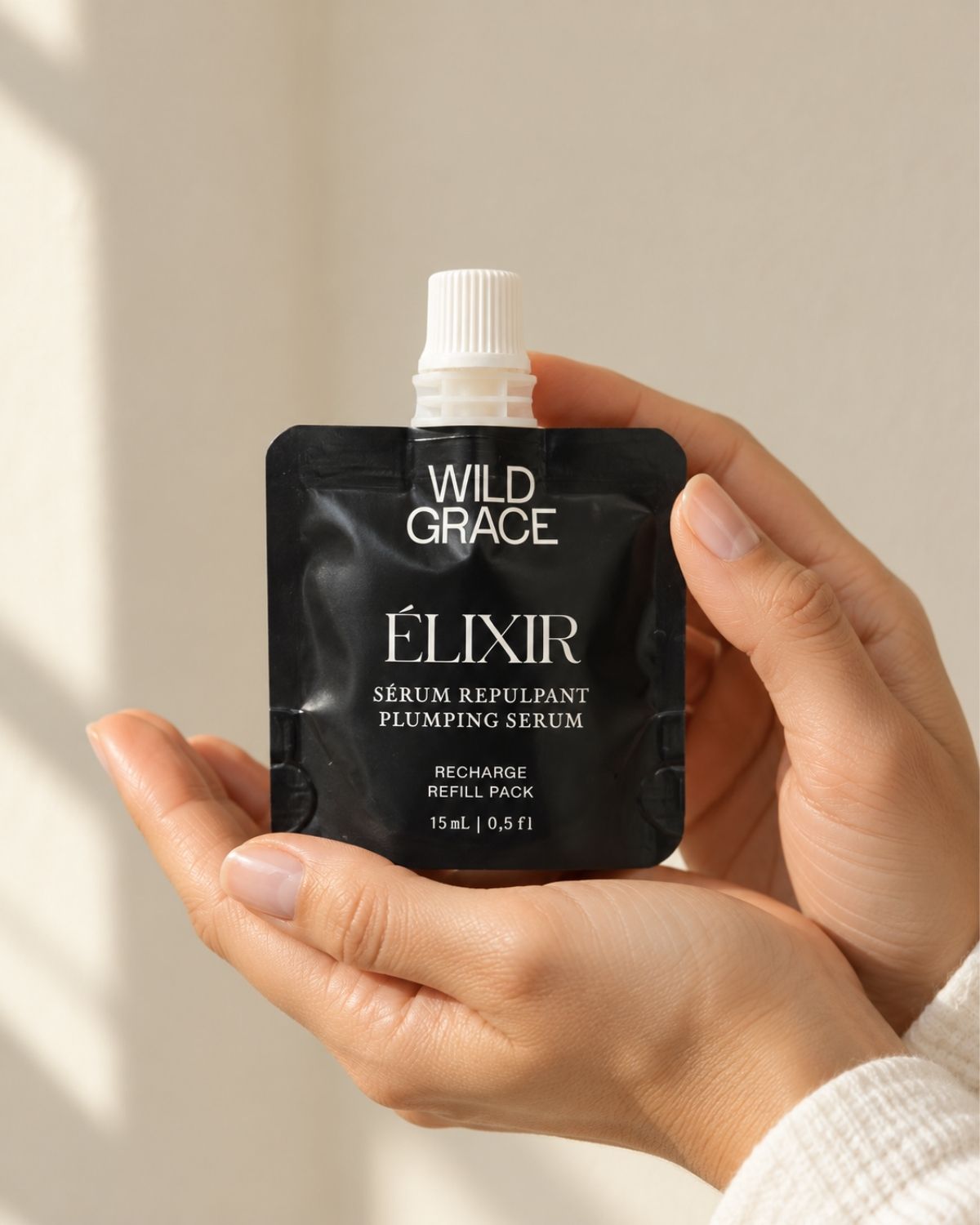 A person's hands holding a black refill pouch of Wild Grace Élixir plumping serum against a warm beige background with soft window shadows.