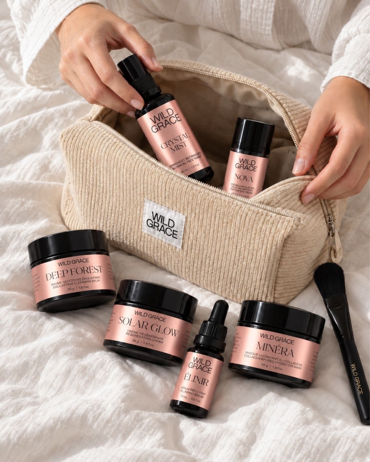 A person's hands open a beige corduroy makeup bag filled with Wild Grace skincare products, including Deep Forest, Solar Glow, Minera, and Clear dropper bottles, with additional products arranged on a white textured blanket.