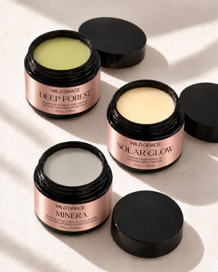 Three open jars of Wild Grace body butters arranged on a light beige surface with soft shadows. The butters are shown in three different shades: a pale yellow-green, a creamy beige, and a white.