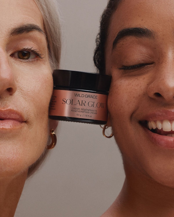 Two women smiling closely beside a jar of Wild Grace Solar Glow Organic Sunscreen Oil, with the product centered between their faces against a soft neutral background.