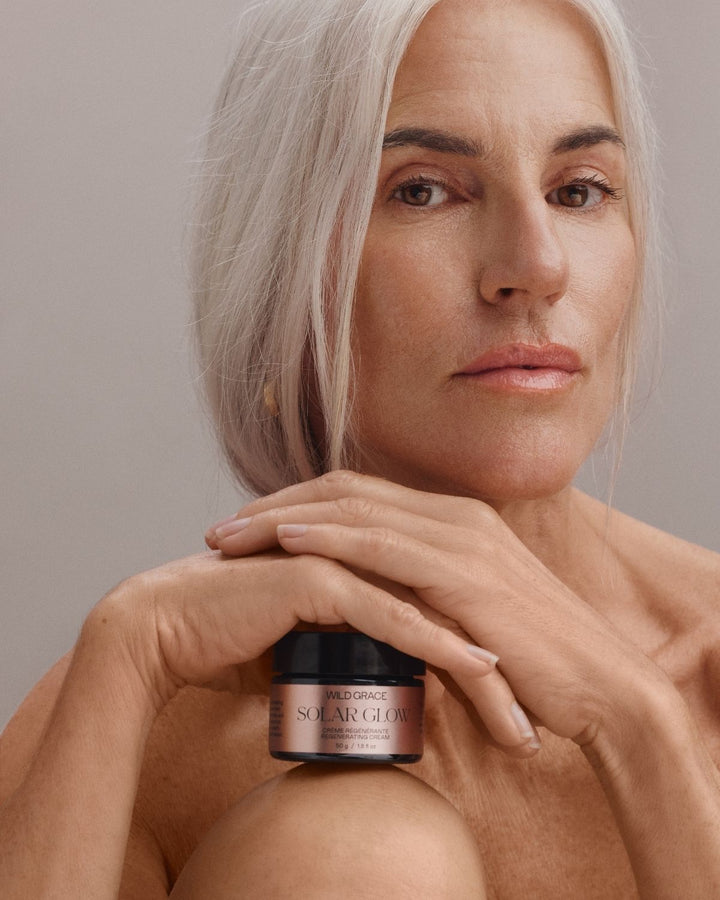 A close-up portrait of a woman with platinum blonde hair holding a small jar of Parfums de Marly Solar Glow Sun Serum against a neutral gray background.