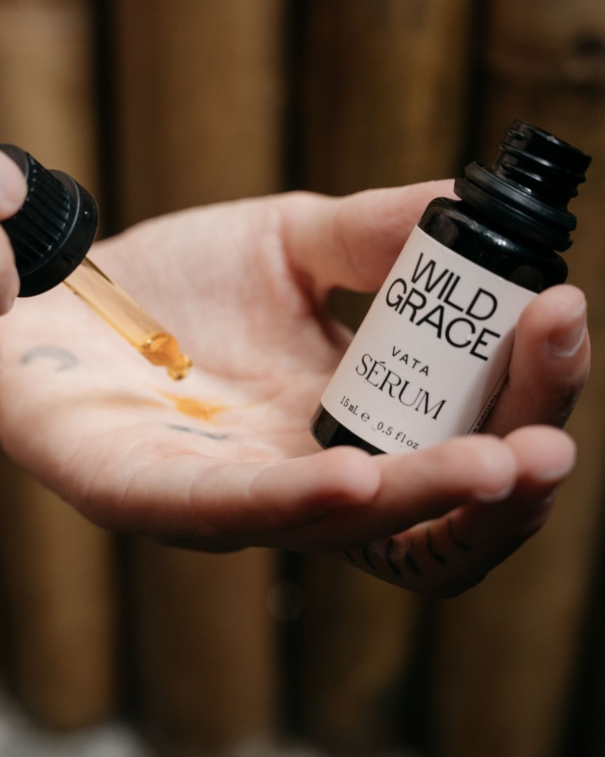 A close-up of a hand holding a small black bottle labeled 'WILD GRACE VATA SERUM' while another hand dispenses amber liquid with a dropper onto the palm, against a blurred wooden background.